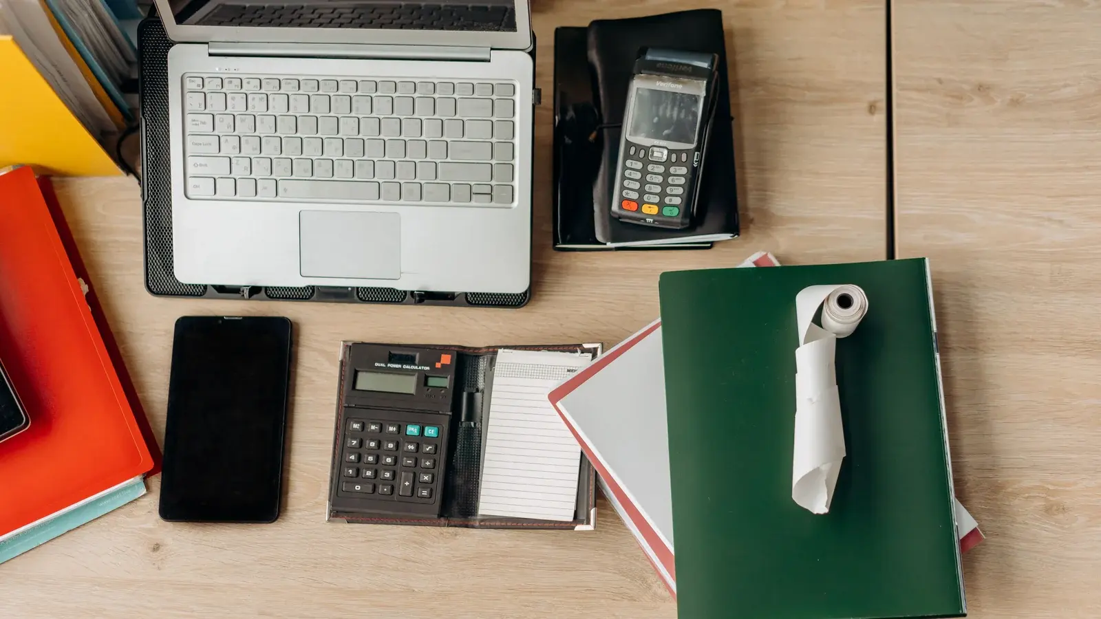 Desk from above with a laptop, calculator, card reader and notebooks